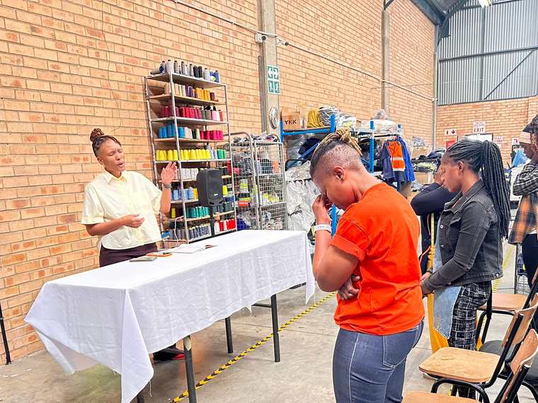 A facilitator standing and speaking at the front of a room while job club members stand quietly at the start of a session.