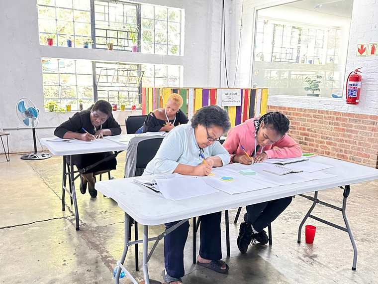 Four Izenzo Job Club members working at tables during a Tuesday workshop, writing in their notebooks.